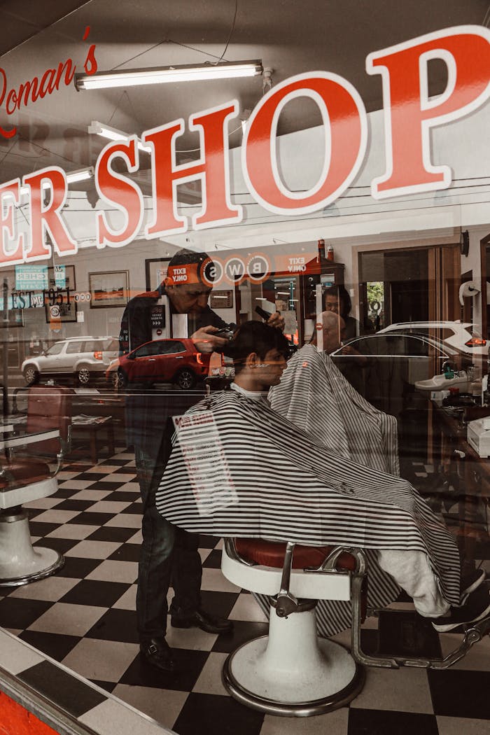 services-01 A barber is giving a haircut inside a classic barbershop, captured through the window.