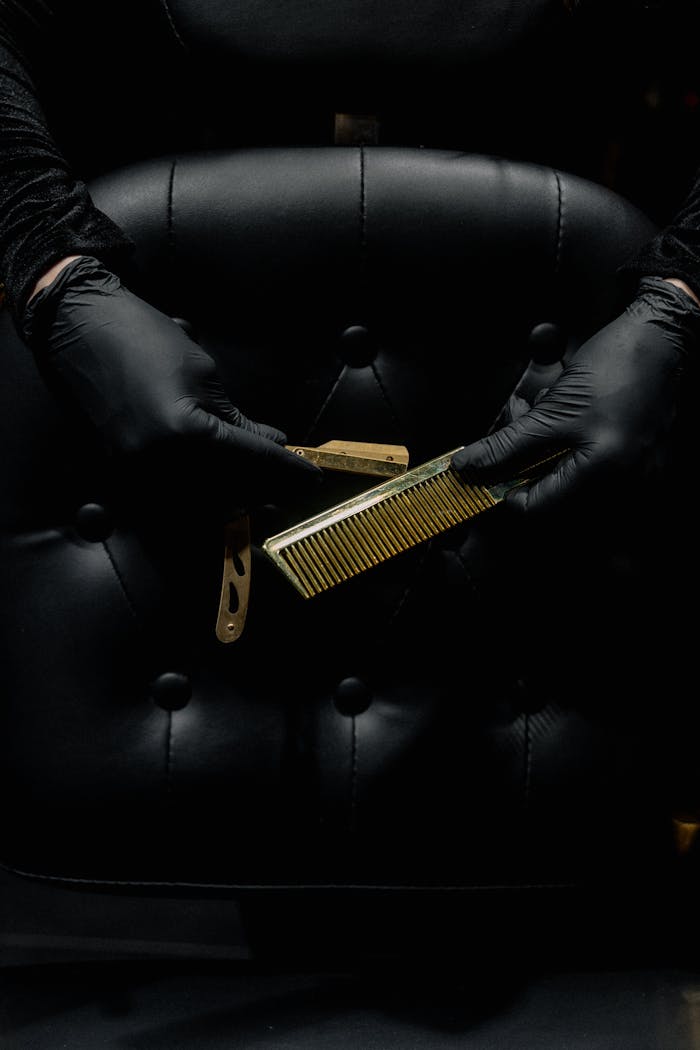 services-04 A barber with gloved hands holding a comb and razor against a leather chair backdrop.
