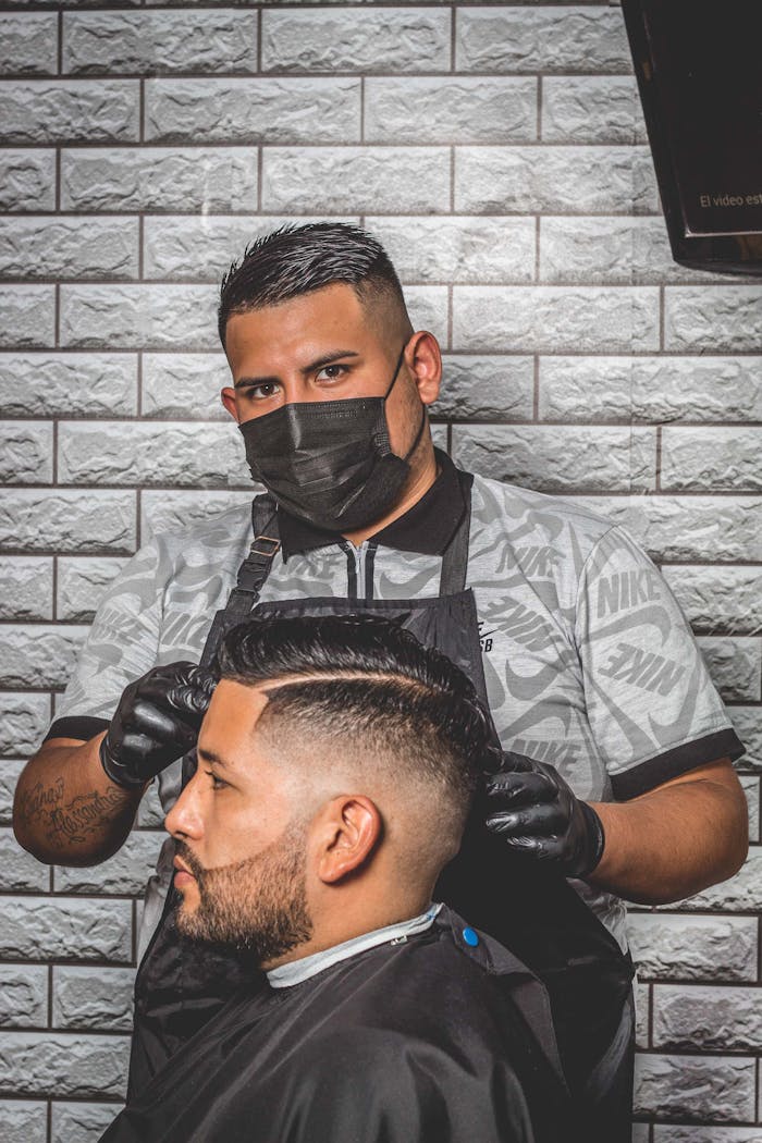 services-02 A barber wearing a face mask styles a client's hair indoors, maintaining safety and style.